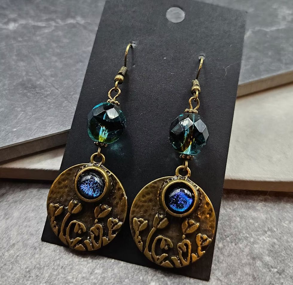 Dark blue and green faceted crystal earrings with a brass tone poppy charm on the bottom that has an inset piece of sapphire blue glass. 