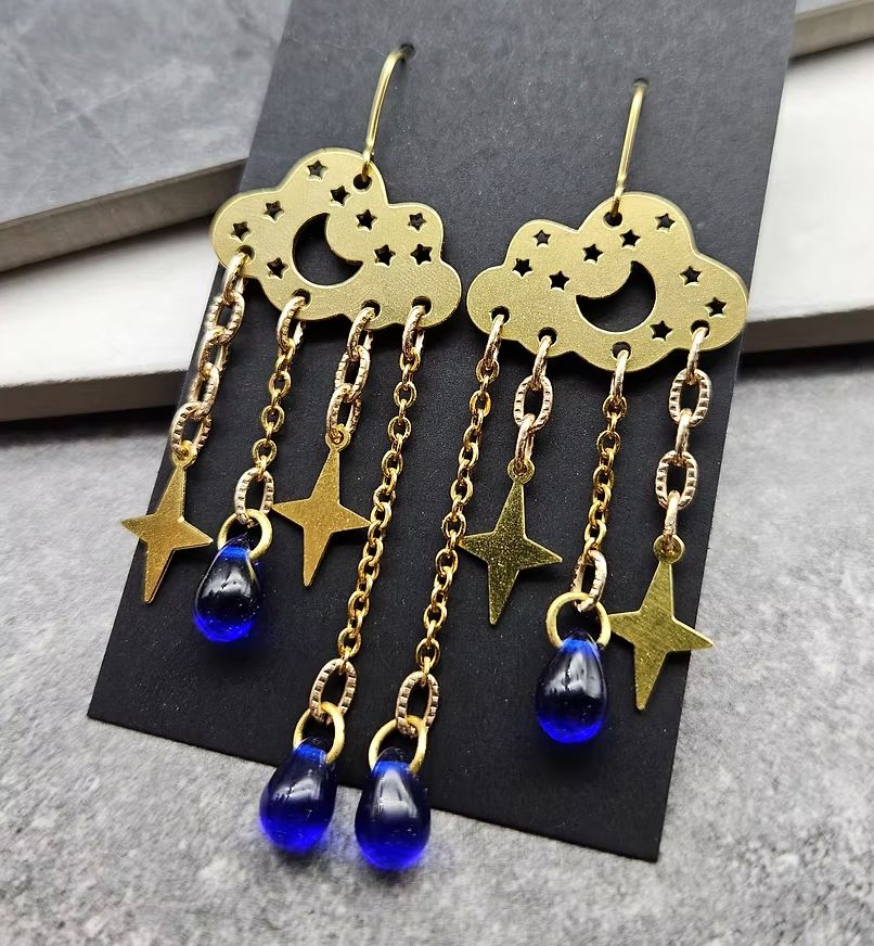 A pair of gold toned chandelier earrings with blue glass drops and gold tone stars hanging from gold toned clouds with a crescent moon and starts cut out 