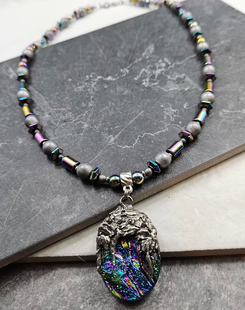 a matte and rainbow henatite necklace that has a dichroic glass pendant in the center with a hand forged metal bail 