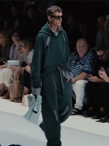 Captura de tela do desfile da Dolce & Gabanna para a coleção Primavera Verão 2026. Na imagem, um modelo masculino veste óculos escuros, um casaco estilo moletom extra-grande com capuz verde escuro com detalhes em verde-claro, uma calça comprida e extra-grande no mesmo esquema de cores. O tecido parece quente.