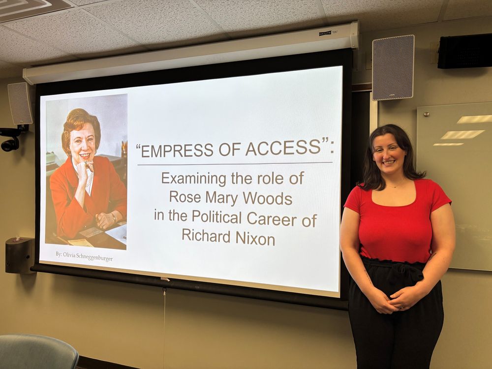 Smiling Schneggenburger in red shirt and dark pants with the title slide of her presentation on Rose Mary Woods, "Empress of Access," to her right.