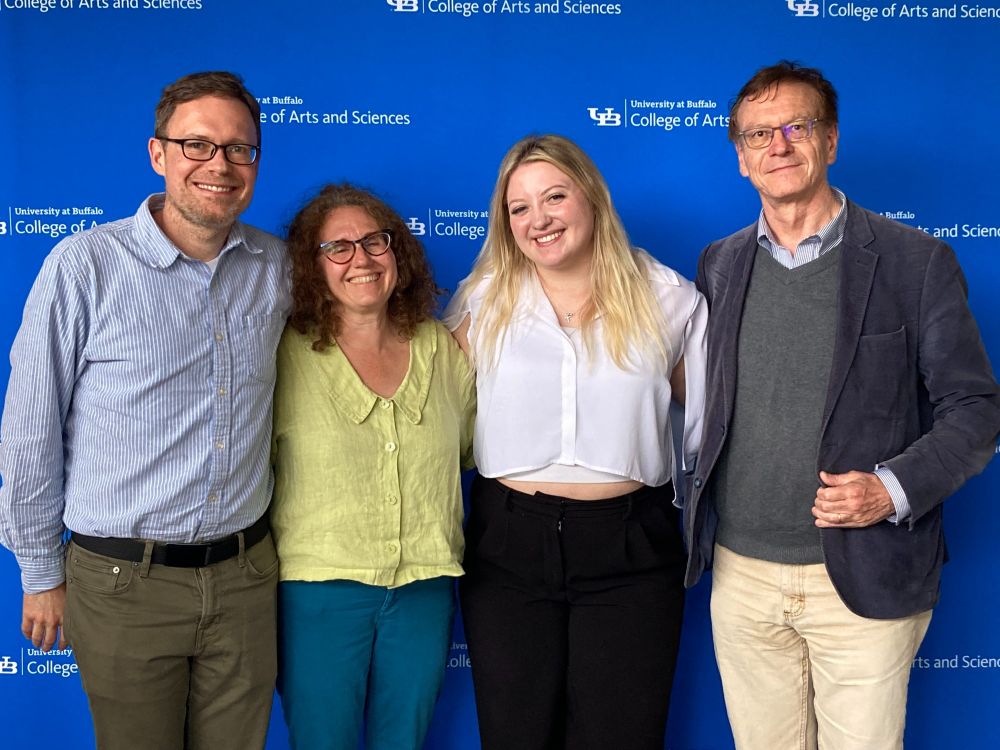 Three light skinned people standing in front of a blue screen. The inner two people are woman and the outer two people are men. they are smiling at the camera. 