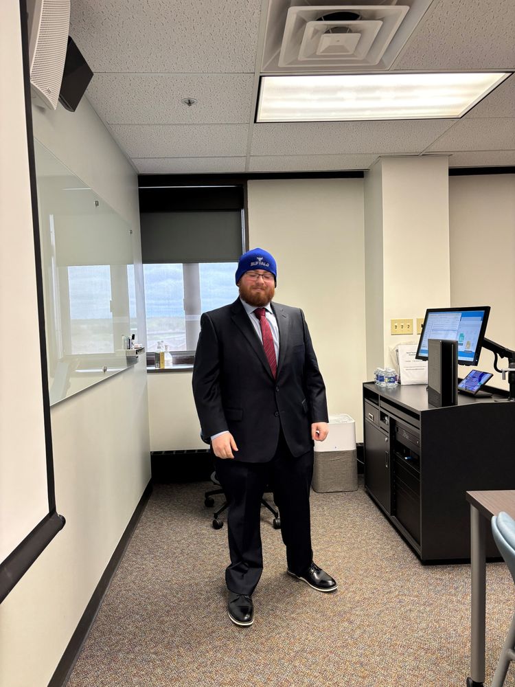 Vener ready to present his work wearing a dark suit and University at Buffalo knit cap with podium to his side and window in the background.