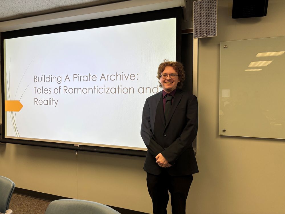 Rechichi, smiling in suit and tie, standing next to his title slide on the screen for his presentation on the pirate archive and his Omeka exhibition.