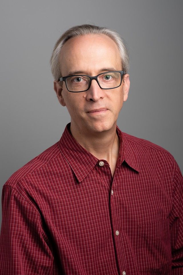 a white man with black glasses and graying hair. he is wearing a checked red button down shirt. 