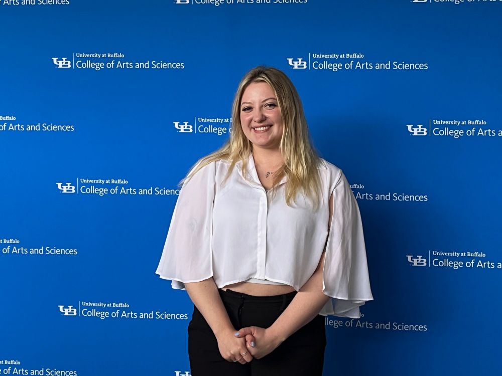 a light skinned woman smiling at the camera in front of a blue screen. the screen reads "University at Buffalo College of Arts and Sciences." She is wearing a white flowing top and black pants. 