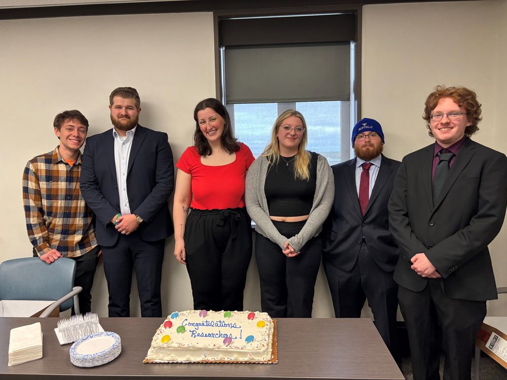 Six smiling students from the Department of History lined up behind the "Congratulations Researchers" white frosted cake with balloons. 