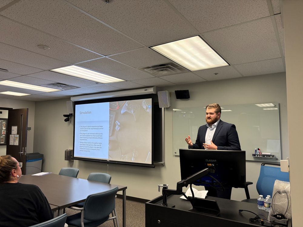 Chandler in blue jacket with light blue dress shirt at podium presenting with a slide on the screen to his right showing the video game set up an describing the war simulation in the games.