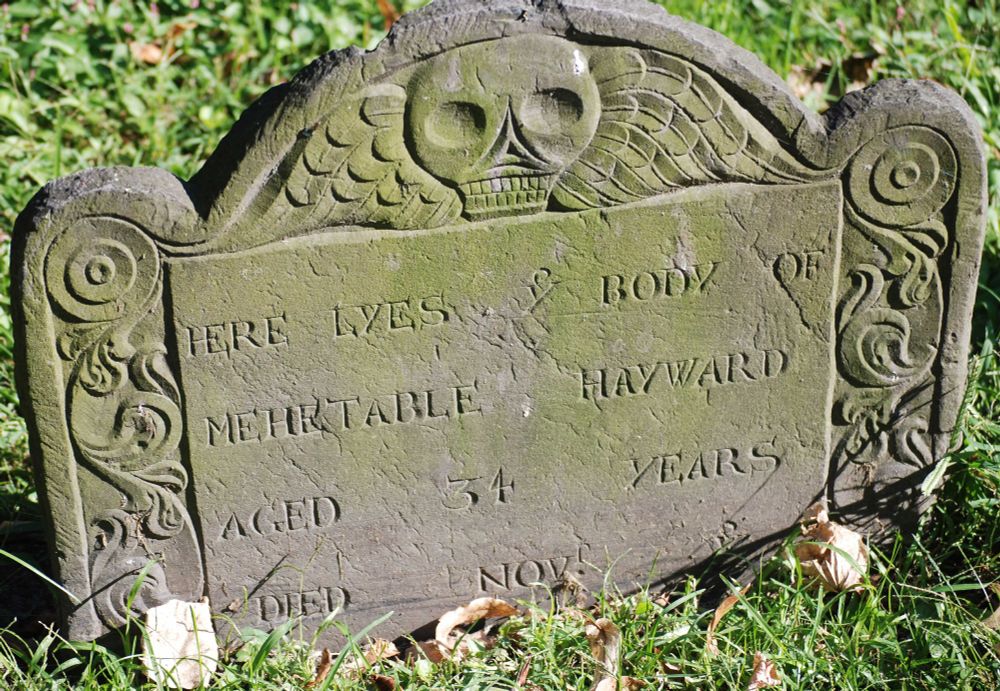 an old Puritan tombstone with a deaths head on the top and scrolls down the sides. in the center the carving reads "Here Lyes the Body of Mehetable Hayward, aged 34 yeras Died Nov " and then the rest of the carving is obscured by the grass. 