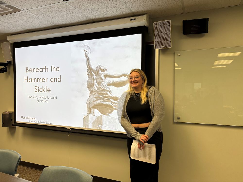 Serrano smiling, wearing a black top and pants with a grey sweater, standing next to her title slide on the screen, reading Beneath the Hammer and Sickle.