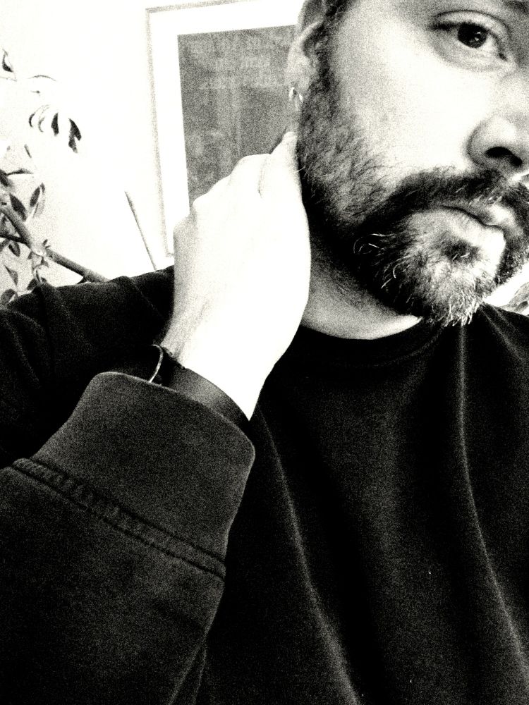 Schwarz weiss Foto, Mann mit Bart und schwarzem pullover greift sich an den Nacken, der Bildausschnitt zeigt nur einen Teil des Gesichts