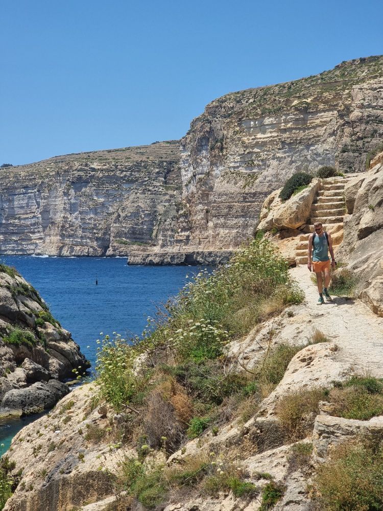 Eine felsige Landschaft mit hohen Klippen, Wasser. Rechts geht ein sommerlich bekleideter mann eine in Stein gehauene Treppe herunter