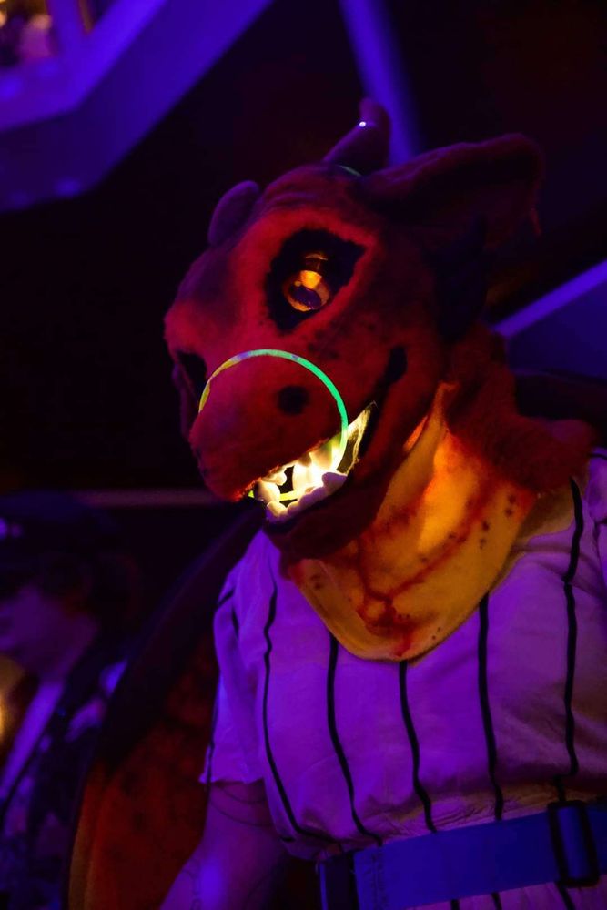 A photo of a person wearing a dragon fursuit, the dragon has reds and oranges, with orange light up eyes. there is glowsticks around the muzzle and horns of the dragon and the mouth is lit up.  The person is wearinga white with thin black stripe play suit.