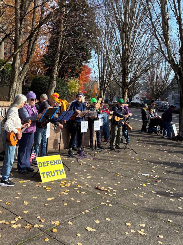 About ten folk with guitars and ukuleles provide musical support  at this weekly protest in northeast Portland, OR. 
  A yellow placard with message, “DEFEND TRUTH”, rests against a music stand,  a small American flag  flying at its corner. 
  Several of the performers have a QR code affixed to their music stands, providing a link to the group’s songbook. 