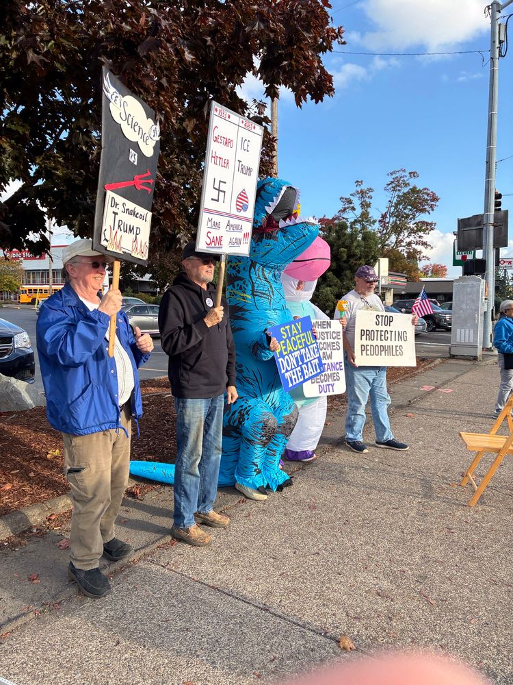 Citizens with protest signs.
From the right: “Science vs Dr Snakeoil Trump”.. sign embellished with flames, and the Devil’s trident 🔥🔱.
  Man with black jacket hold sign which compares 1933 Gestapo and Hitler to 2025 ICE and Trump..includes entreaty, “Make America Sane Again ‼️”.
  A blue T-Rex hold sign reminding us, “Stay peaceful, don’t take the bait”.
  A white unicorn bears a placard, with message, “when injustice becomes law resistance becomes duty”. 
  At far right, citizen holds a small American flag, & a sign which reads, “STOP PROTECTING PEDOPHILES”