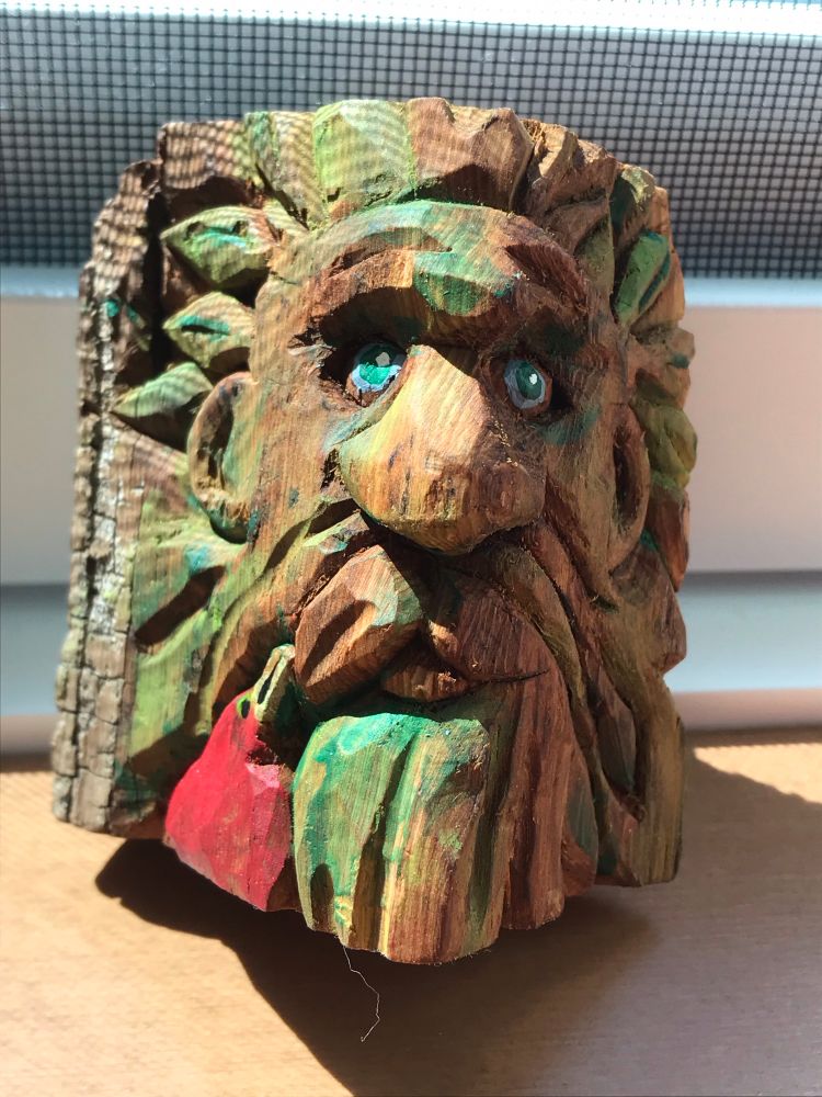 A carved wood-spirit. Aprx 3” tall, hewn from cottonwood bark. His face is framed by green leaves- he’s accompanied by a small red bird at his lower left.