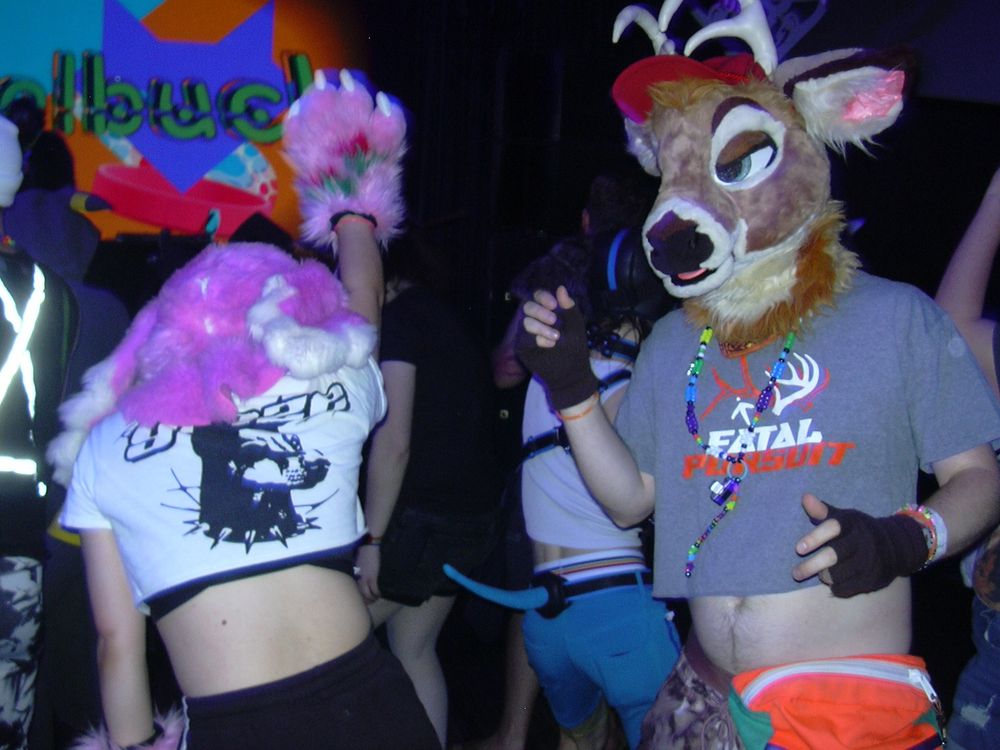A digicam flash photo of Theory the pink dragon in partial fursuit, dancing with one arm up and facing away from the camera. Adjacent, Atmus the deer dances in partial fursuit, facing toward the camera with a half-lidded gaze. His colorful kandi necklace jingles with his movements. The crowd bounces around them, and in the background the screen above the stage reads “russelbuck” with colorful graphics.