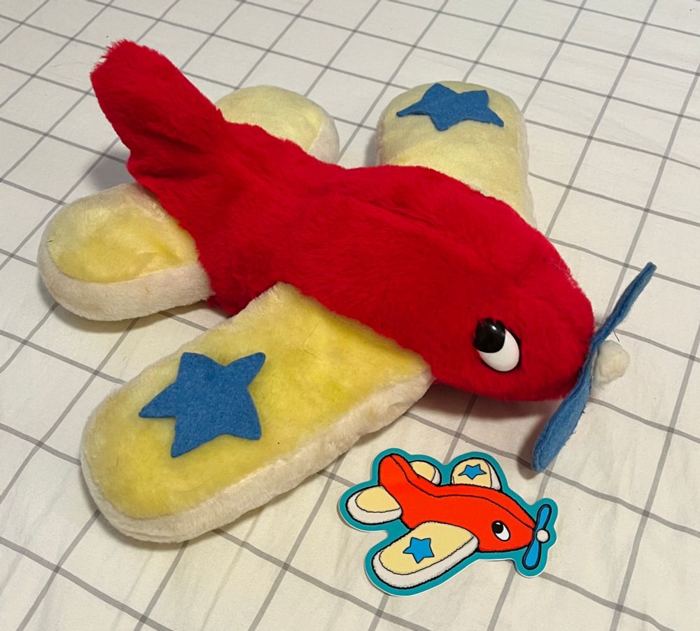 A photo of a cutesy plush airplane resting on a grid pattern sheet. It has a red body, yellow wings, and a blue propeller nose. It has a blue felt star on each wing. It’s plastic eyes sparkle innocently. Next to the plush, there is a sticker featuring a drawing of the airplane. 