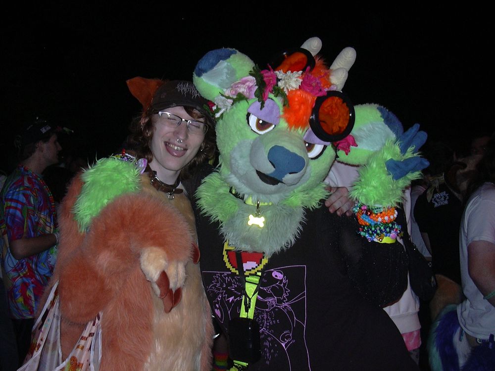 Digicam flash photo of Bandit/Pipsqueak the squirrel and Russelbuck the green deer. Pipsqueak is wearing his bodysuit and paws but no fursuit head, sticking his tongue out playfully. Russy is in their head and fursuit hands, flashing a peace sign. 