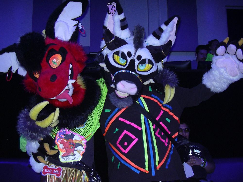 A digicam flash photo of two partial fursuiters posing together. On the left is a red dragon character named Mordecai, wearing a neon green mesh crop top, a shiny gold hip bag, and glowstick bracelets as fursuit ear jewelry. On the right is a black and white canine character with horns, wearing a black top with colorful UV patterns, and kandi jewelry around their muzzle and ears.