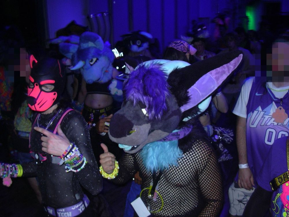 A digicam flash photo of the crowd at PLUSH dance party. In the foreground, a  purple dragon partial with blue horns, wearing a fishnet top. Adjacent, a pup in a neon pink neoprene hood, sheer top, and bulldog style harness. More bodies and fursuit heads move in the background. 