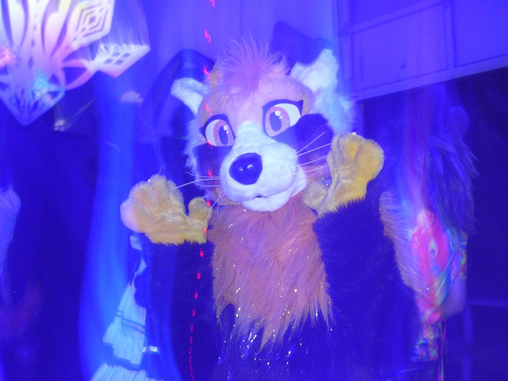 A digicam flash photo of a fursuit resembling a red panda with floppy ears andwhite whiskers. Their yellow hand paws are raised for the picture. The light is distorted, creating a wispy blue haze over the photo, and a repeated red blip trails through the center in a vertical dotted line. 