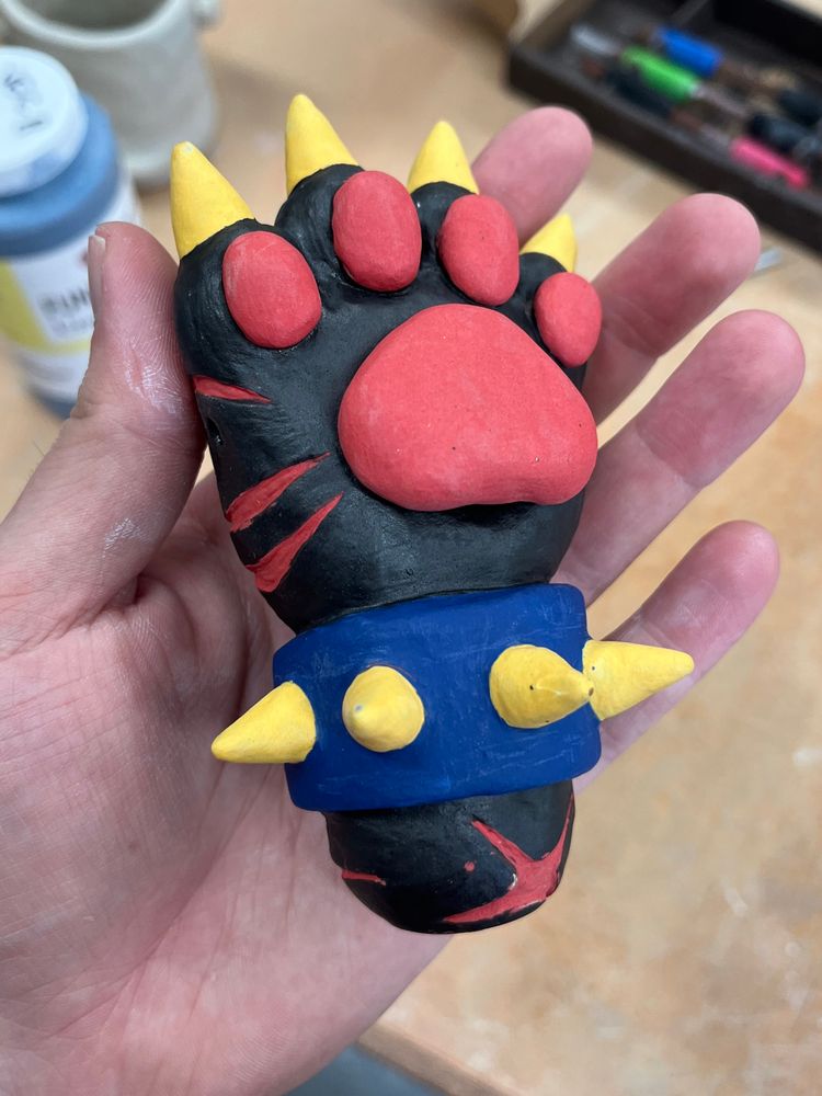 A photo of a ceramic pipe made to look like a black paw with yellow claws, red paw pads, and red scars. It is wearing a blue cuff with yellow spikes, and it has a matte finish. The central paw pad is a separate piece, serving as a lid for the bowl.