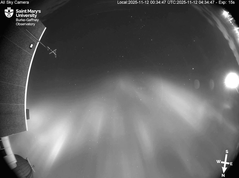 A still frame from the video above at 12:34AM AST. The black and white image shows the wavy band of aurora through the middle of the field of view, all the way from left to right