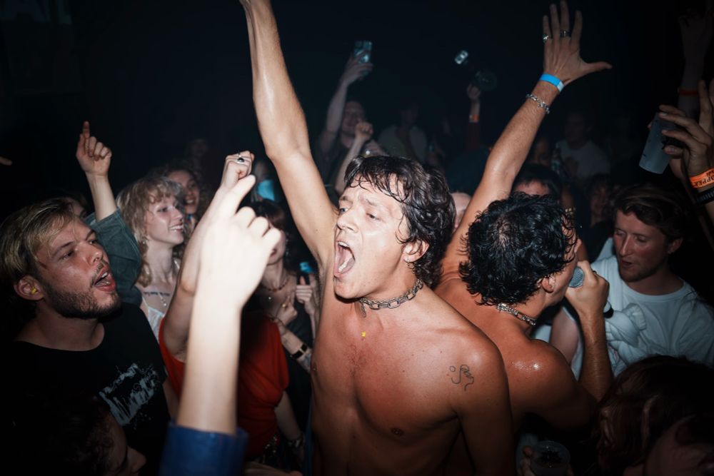 A photo of Faux Real playing, the two brothers are topless in the audience with their arms raised up and their mouths wide open signing. The audience around them looks excited and is pressed together tightly.