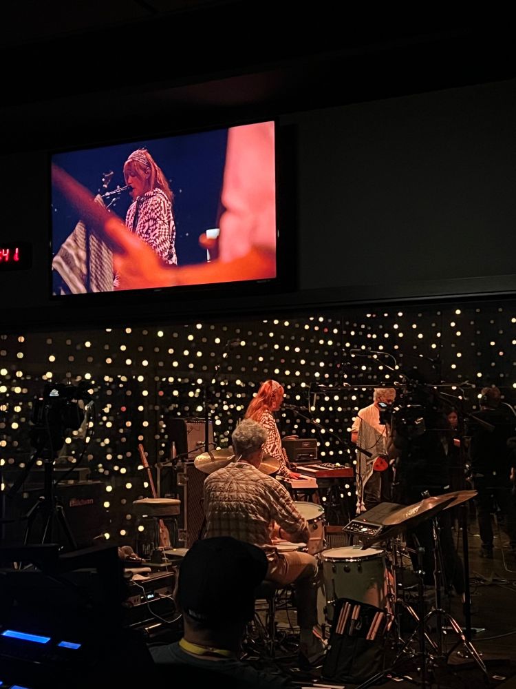 Blonde Redhead performs in KEXP’s studio. Photo is taken from the perspective of a room looking into the studio through a pane of glass. A live feed of the performance is shown above the glass, on a tv hanging on the wall.