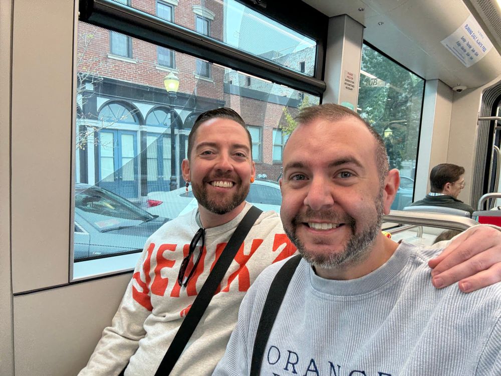 Nate and William, two white men, take a selfie while seated on a streetcar.