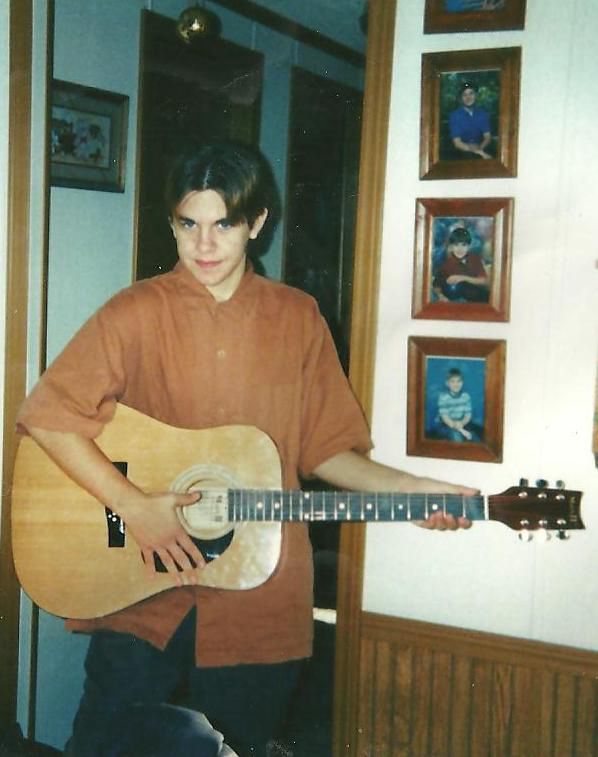 Me as a teen dressed in an over sized brown shirt and holding a guitar. My hair styled with a middle part.
