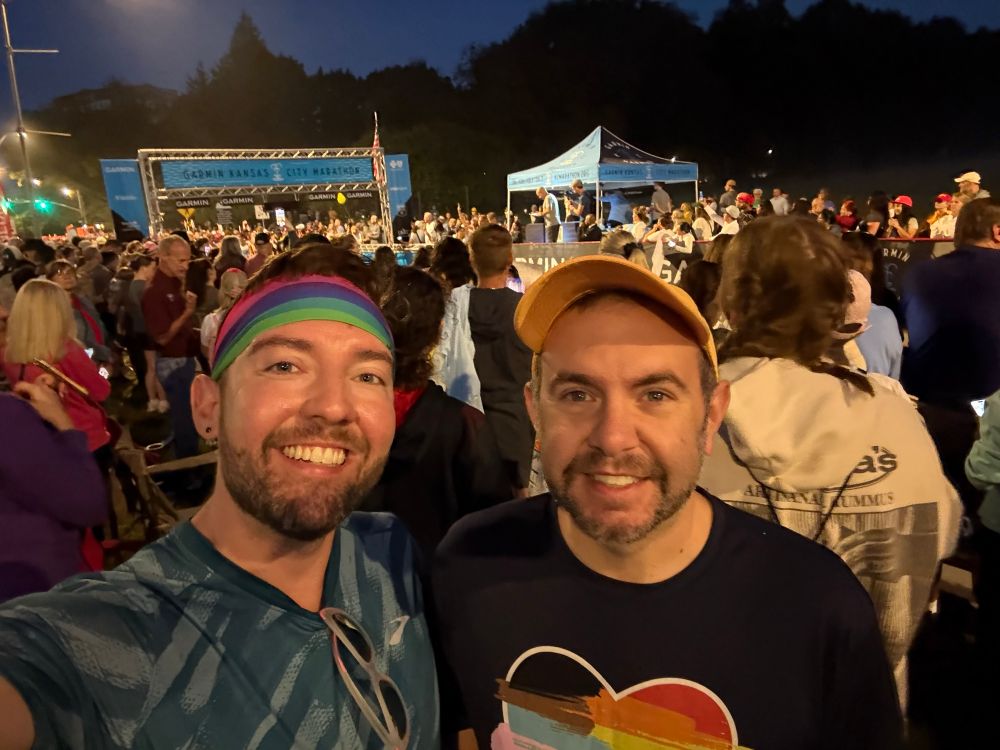 Two white men taking a selfie in front of a crowd of people, an announcer tent, and a run start line 