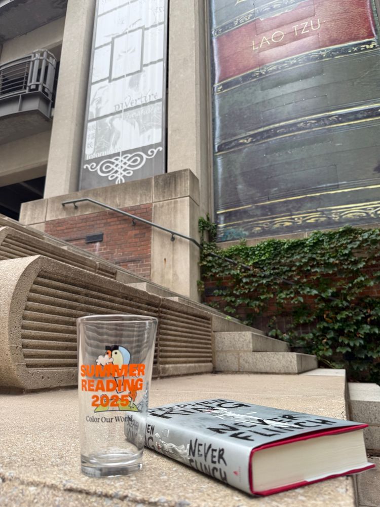 A pint glass next to a book by Stephen King on the stone steps to a parking garage entrance. The stonework is shaped like books.