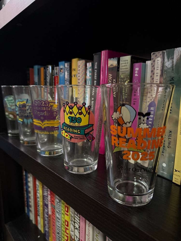 Five decorated pint glasses on a book shelf with books behind them. The closest glass has a hot air balloon and “Summer Reading 2025” printed in orange text on it.