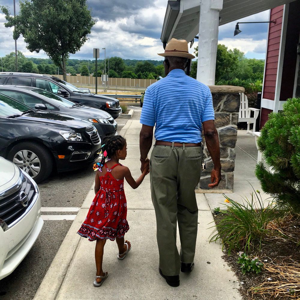 Image of a young girl in a red dress holding hands with her great grandfather as they walk on a sidewalk. 