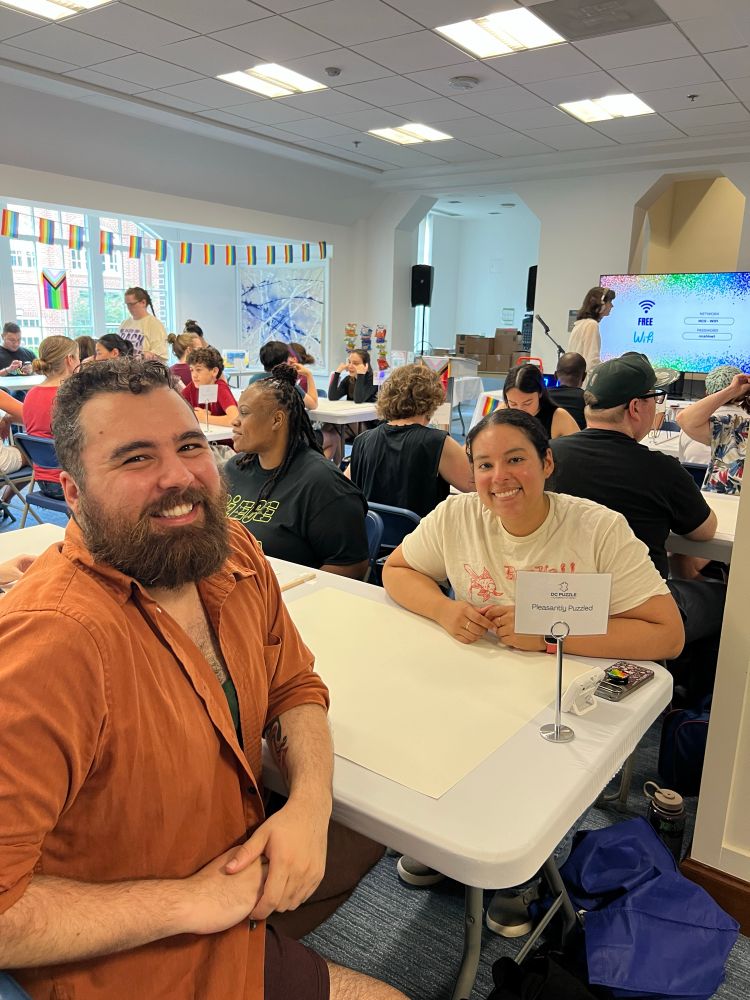 A man and a woman smiling at the camera, seated at a table in a crowded room full of other tables. A small sign on their table shows their team name is “Pleasantly Puzzled.” 