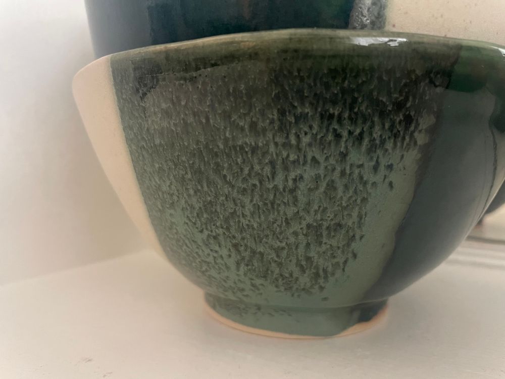 Close up of the side of a bowl showing the eggshell glaze to the left, dark green glaze to the right and flowy combo of the two in the center that shows up as organic pattern of dark green feathering over light green background. 