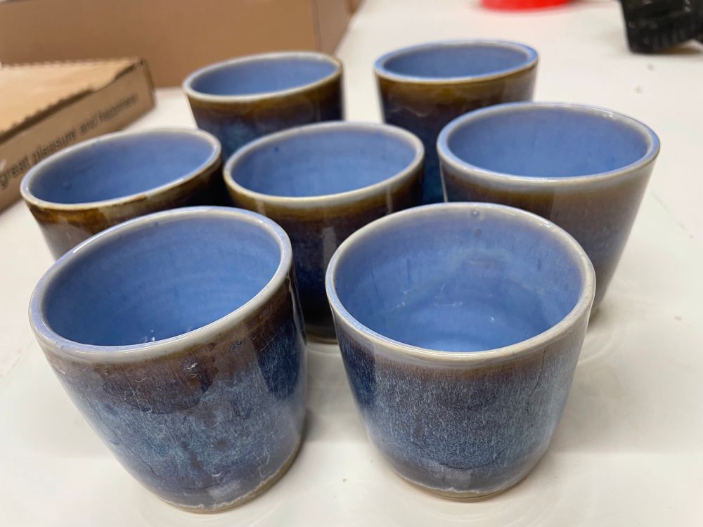 7 cups on a white table. The cups are blue on the inside and dark reddish brown with intense blue speckles all over. 