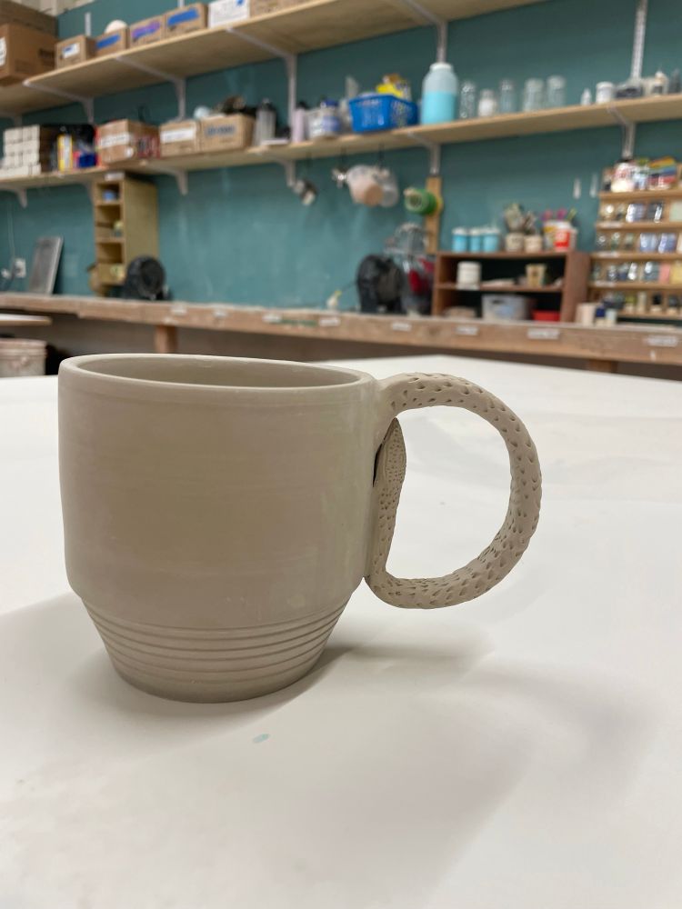 Greenware mug with tapered bottom 1/3 and sculpted handle sitting on a white table
