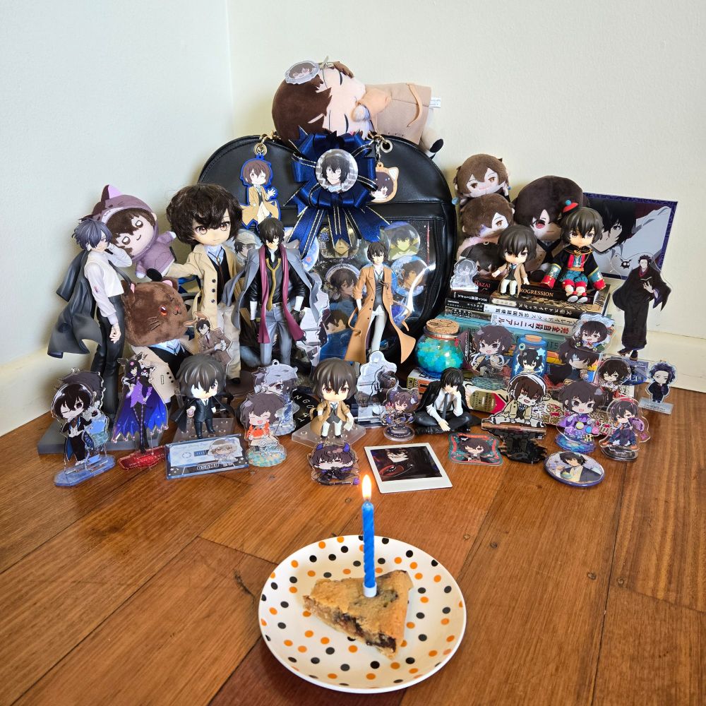 Picture of my collection of dazai items. Pictured is an ita bag full of charms, plushies, figures, and standees. In the foreground is a small plate with a slice of cookie with a candle in it. 