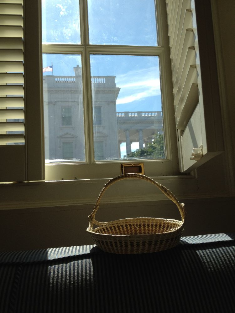 A Charleston sweetgrass basket made by an African American woman of Gullah Geechee descent sits in the late day sunlight streaming in through a window with a view of the North Portico of the White House