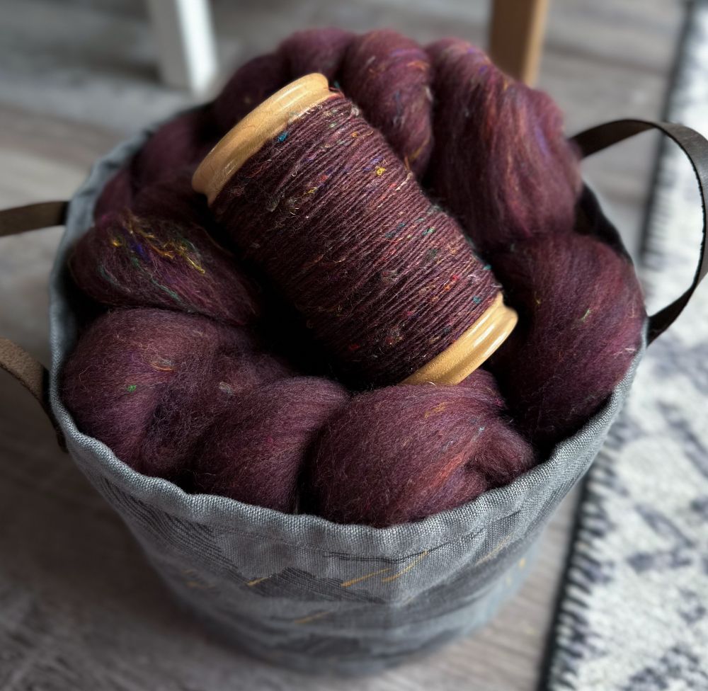 A gray fabric bucket sits on the floor containing dark red carded fiber. On top of the fiber is a full bobbin of the spun fiber.