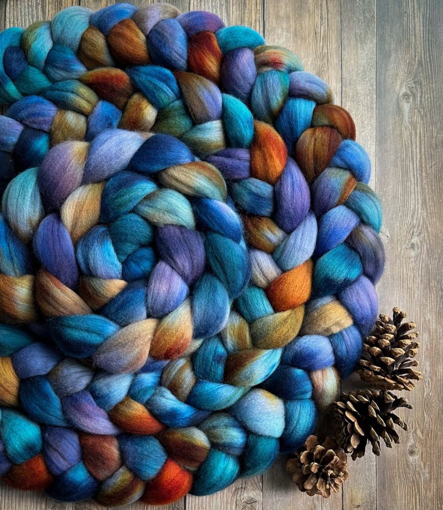 Braided Polwarth wool sits on a wooden backdrop. The wool has been dyed in shades of blue, light purple, brown, and orange. Three pine cones sit next to the wool.