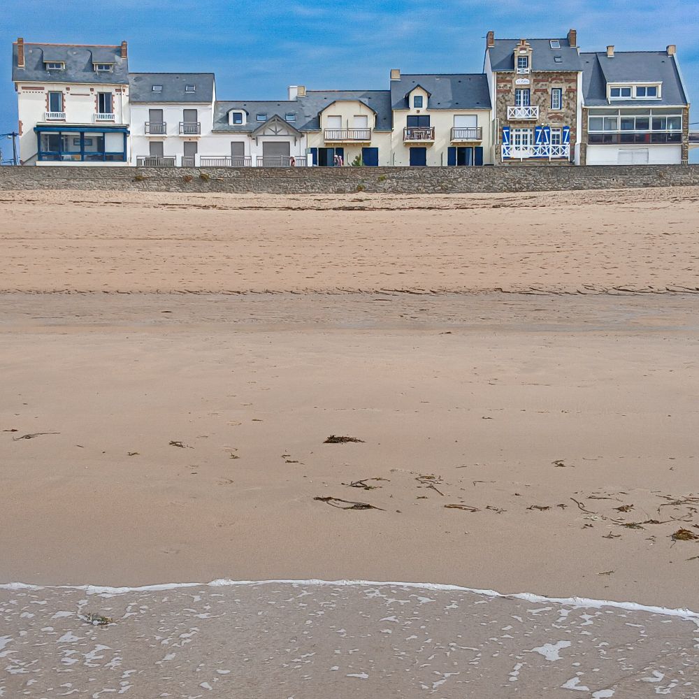 Am oberen Bildrand blauer Himmel, darunter eine Häuserreihe, eine Natursteinmauer und der Strand, der den größten Teil des Bildes einnimmt. Am unteren Bildrand ein bisschen Wasser mit Gischt.
