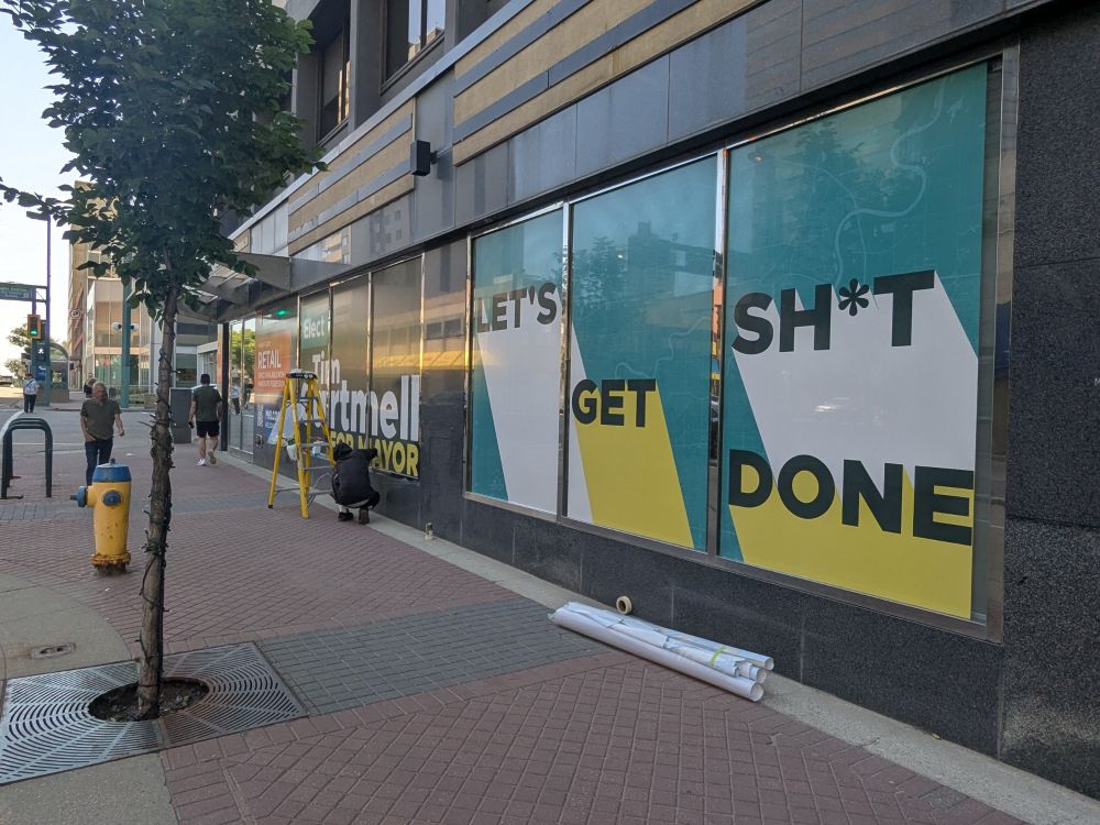 Workers apply vinyl wrap to the windows of an office building advertising the campaign of Tim Cartmell for mayor of Edmonton. The slogan is LET'S GET SH*T DONE.