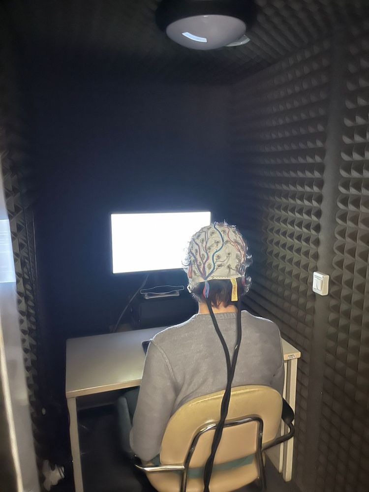 Adult participant with an EEG cap on in front of a computer screen in a sound-proof testing booth