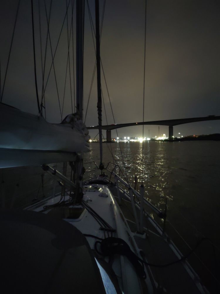 A photo taken from a yacht deck, its dark with a few clouds in the sky. In the distance over the bow are the lights of Ipswich through the Orwell bridge.