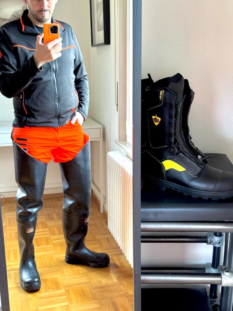 Man in a mirror, wearing high black rubber waders, orange high viz pants and a black sweater. Firefighters boots are visible as well. 