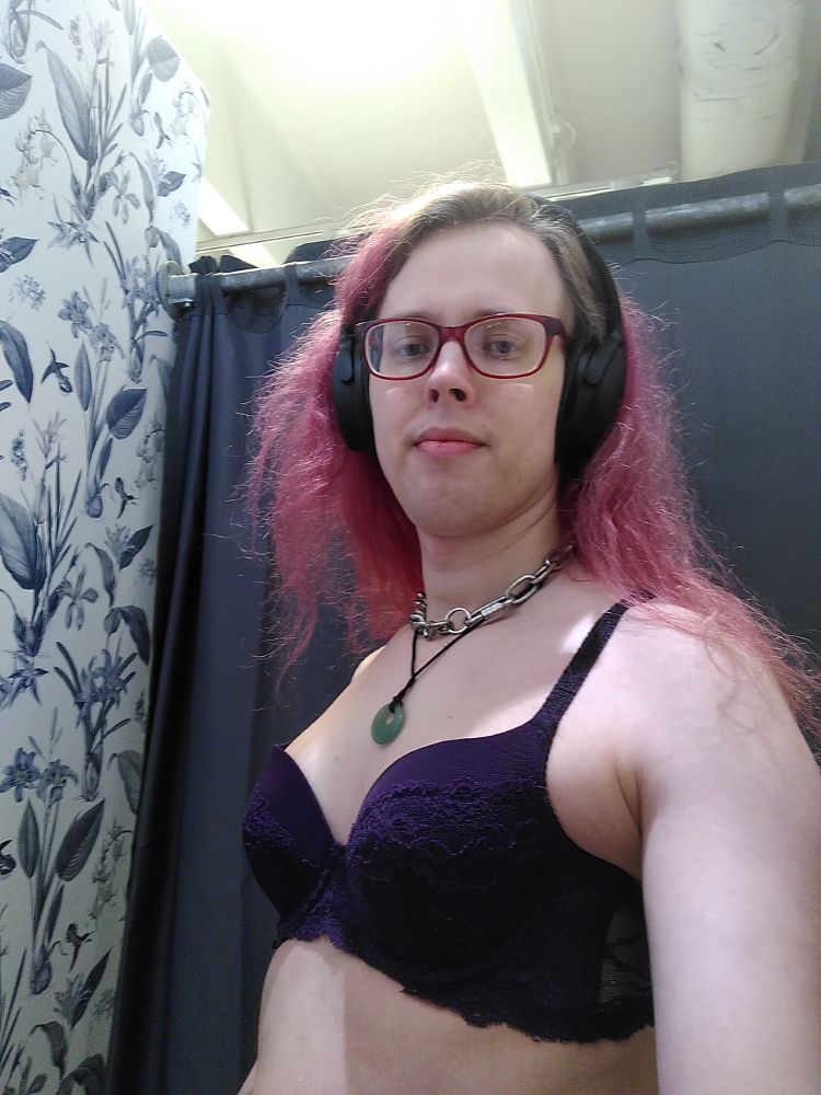 Me a transfemme lady, red loo messy hair with lighter har showing at the roots, I’m taking a selfie in the changing room trying out the brah before buying it, only upper body from just above the navel showing in picture, I’m wearing  my black headphones, I have a medium heavy chain necklace and my jade pendant on a string hanging around my neck I’m wearing a dark purple lace patterned brah with a small part showing of see through fabric in the back part of the brah